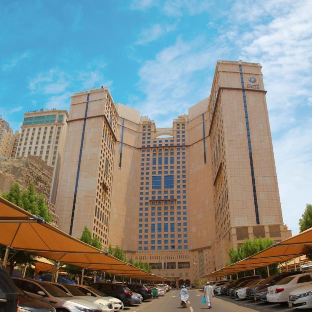 Tall hotel buildings with arched facade under a blue sky, surrounded by parked cars and trees.