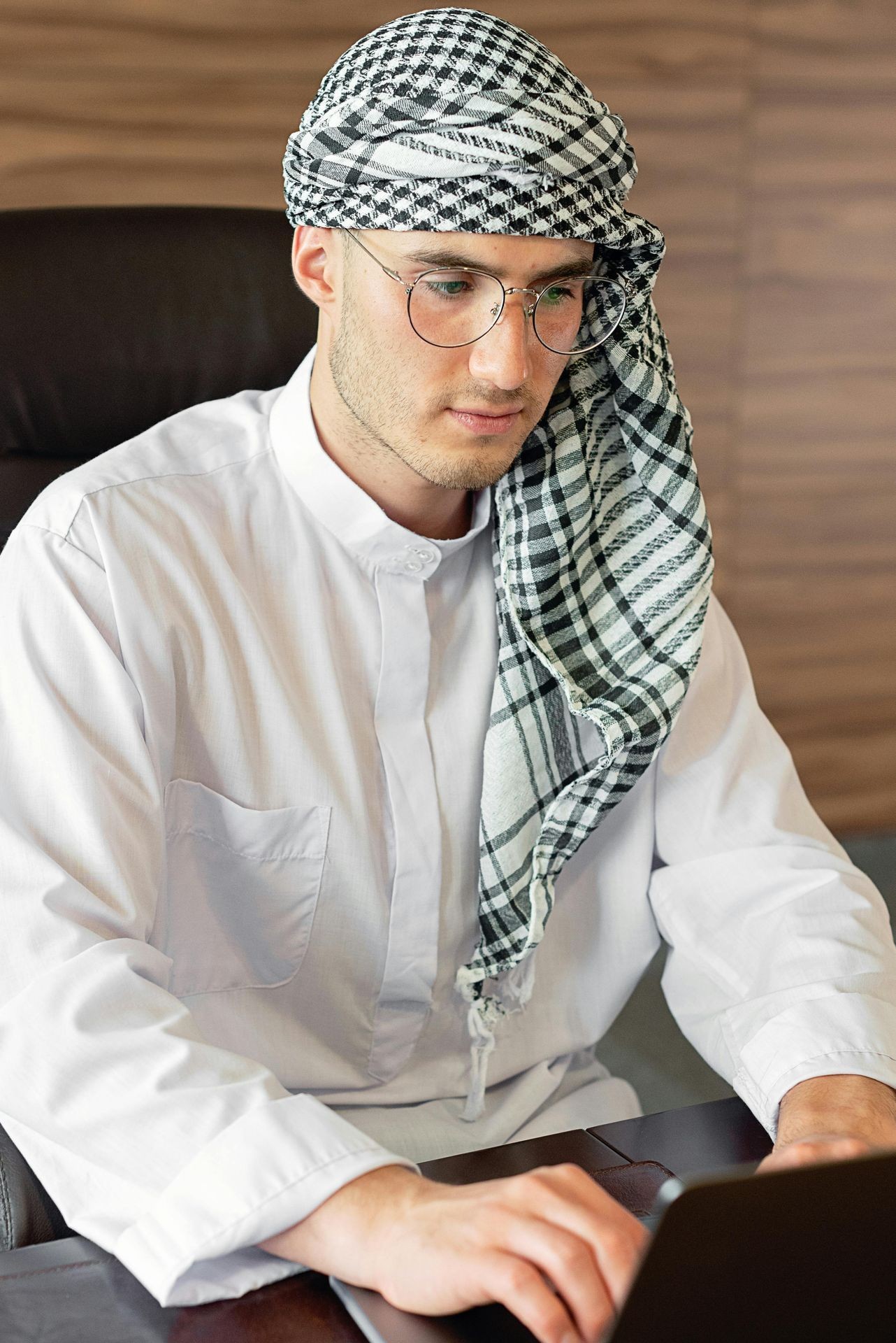 Man in traditional attire and glasses focused on a laptop at a desk.
