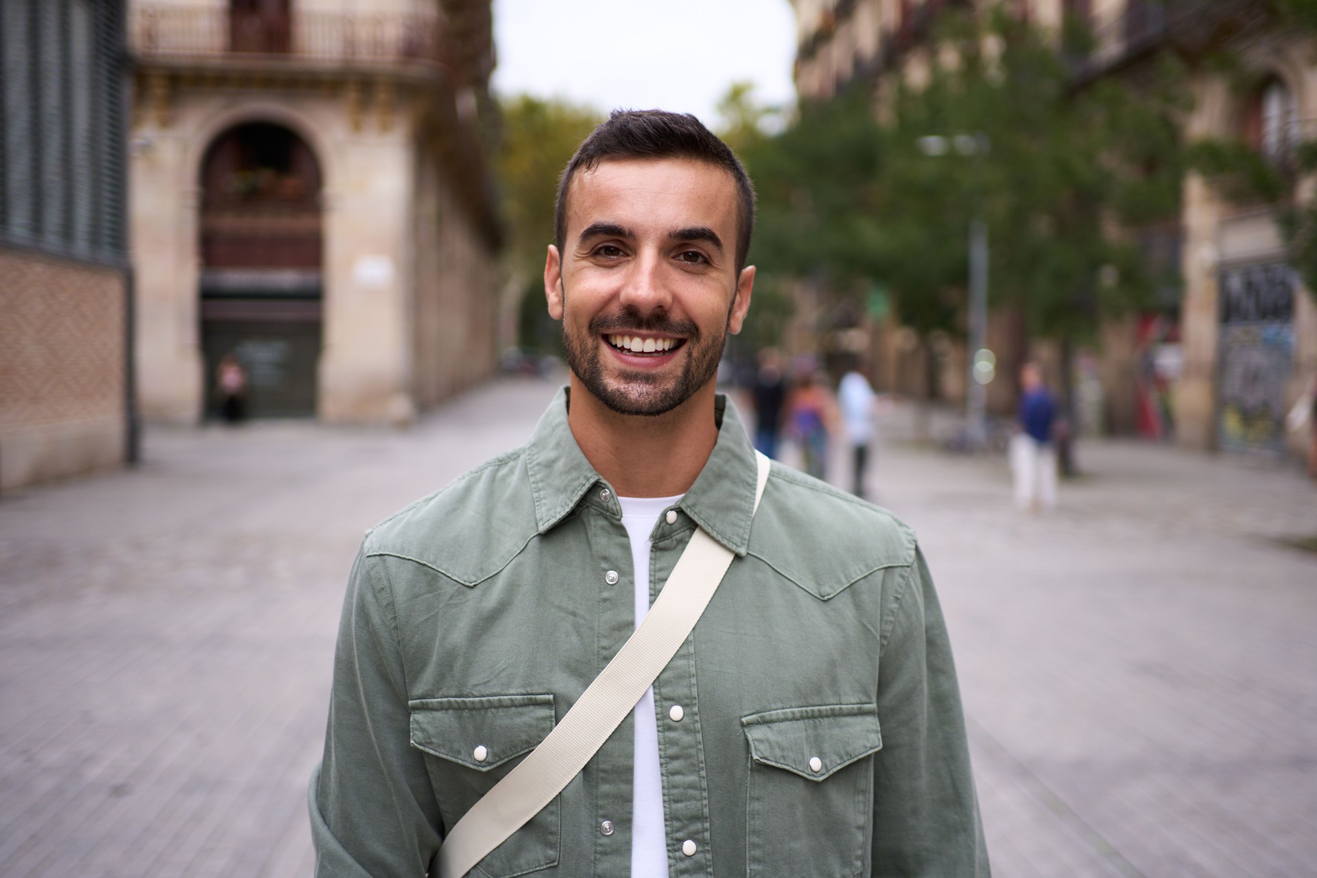 Portrait young bearded handsome man stands looking at camera smiling on street in city of Barcelona.